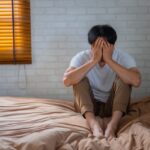 朝ベッドの端に腰かけて頭を抱える男性