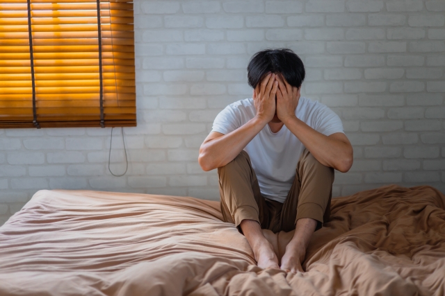朝ベッドの端に腰かけて頭を抱える男性